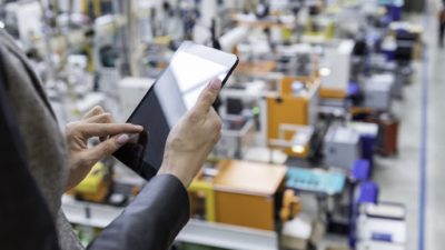 Horizontal color image of businesswoman - unrecognizable person - working with digital tablet in large futuristic factory. Woman standing on top of a balcony, holding touchpad and checking inventory of a manufacturing company on touchscreen tablet. Focus on businesswoman's hands holding black tablet, futuristic machines in background.